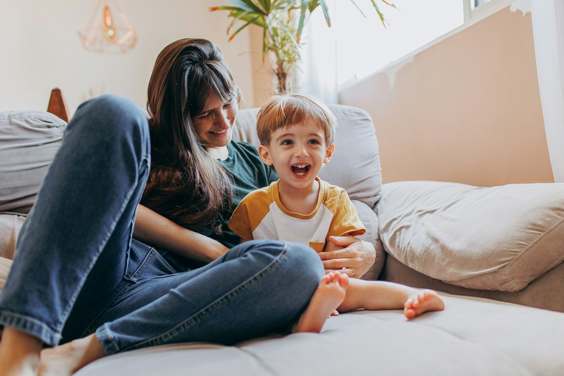 Eine Mutter sitzt mit ihrem lachenden Sohn auf dem Sofa in einem hellen Wohnzimmer